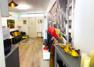 Una moderna sala de estar con suelos de madera clara, un sofá gris, detalles en amarillo y el arte mural de un coche antiguo dan vida al estilo Bluebenalmadena. Un modelo de scooter amarillo en la estantería añade encanto a este espacio luminoso y naturalmente iluminado.