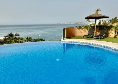 Una piscina infinita redonda en Bluebenalmadena da al océano con aguas azules cristalinas. Al fondo, palmeras, tumbonas y una sombrilla de paja en un patio ofrecen una serena vista de la costa bajo un cielo despejado.