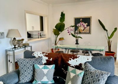 Un salón moderno con un sofá gris decorado con cojines estampados y una manta de piel de vaca. Al fondo, el estilo Bluebenalmadena brilla a través de una mesa de comedor de cristal, plantas de interior y un cuadro enmarcado en la pared.