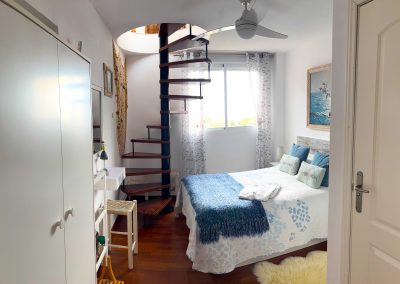 Un acogedor dormitorio Bluebenalmadena con una escalera de caracol, una cama doble con ropa de cama azul y blanca, una ventana con cortinas transparentes, un armario blanco, tocador y ventilador de techo. La luz natural ilumina el espacio.