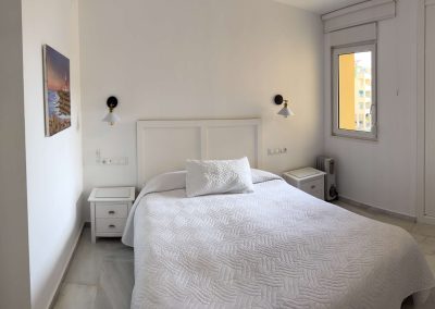 Un dormitorio luminoso y minimalista de Bluebenalmadena con una cama de matrimonio, ropa de cama blanca, dos mesillas de noche, lámparas de pared, una ventana iluminada por el sol y un armario. Una foto enmarcada cuelga de la pared izquierda.