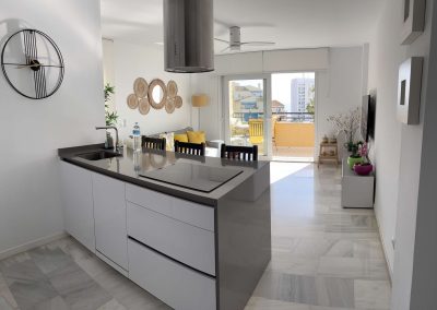 Moderna y luminosa cocina abierta y sala de estar con armarios blancos, encimera gris, reloj de pared, mesa de comedor y detalles amarillos. Las puertas correderas se abren a un soleado balcón Bluebenalmadena con asientos al aire libre.