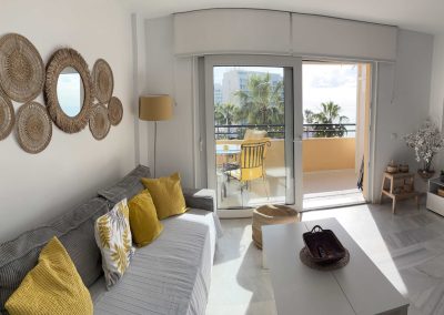 Luminoso salón con un sofá gris, almohadas amarillas y decoración tejida en la pared, que da a un balcón con una silla y una mesa amarillas. Los grandes ventanales de Bluebenalmadena dejan entrar la luz del sol y ofrecen vistas a las palmeras y al mar.