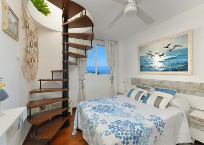 Un acogedor dormitorio Bluebenalmadena con una escalera de caracol, ropa de cama con motivos azules, ventilador de techo, ventana con vistas al océano y un gran cuadro de gaviotas sobre el cabecero.