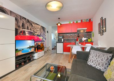 Un moderno estudio de Bluebenalmadena con una cocina roja, un mural en blanco y negro de coches clásicos, un sofá gris, una mesa de centro de cristal y un televisor que muestra una escena de billar. El espacio está muy iluminado y decorado con estilo.