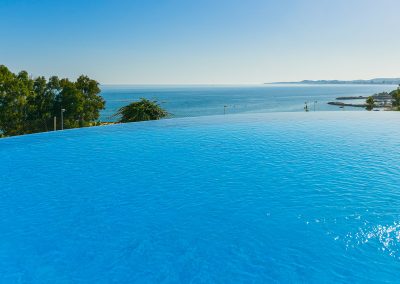 Piscina infinita de aguas cristalinas con vistas a un océano en calma, con la costa lejana y el cielo azul visibles en el horizonte. Los árboles y la vegetación bordean las vistas de Bluebenalmadena, creando una tranquila escapada costera.