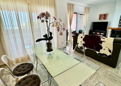 El luminoso y moderno salón-comedor de Bluebenalmadena cuenta con suelos de mármol, una mesa de comedor de cristal con cuatro sillas transparentes, un centro de mesa con una orquídea en maceta, cortinas beige y un sofá con una manta de piel de vaca frente al televisor.