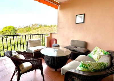 Un acogedor balcón de Bluebenalmadena con sillas de mimbre, una mesa redonda de cristal, un banco acolchado y cojines de hojas verdes. Las baldosas de terracota y una pared melocotón añaden calidez, mientras que un toldo naranja ofrece relajantes vistas de la vegetación exterior.